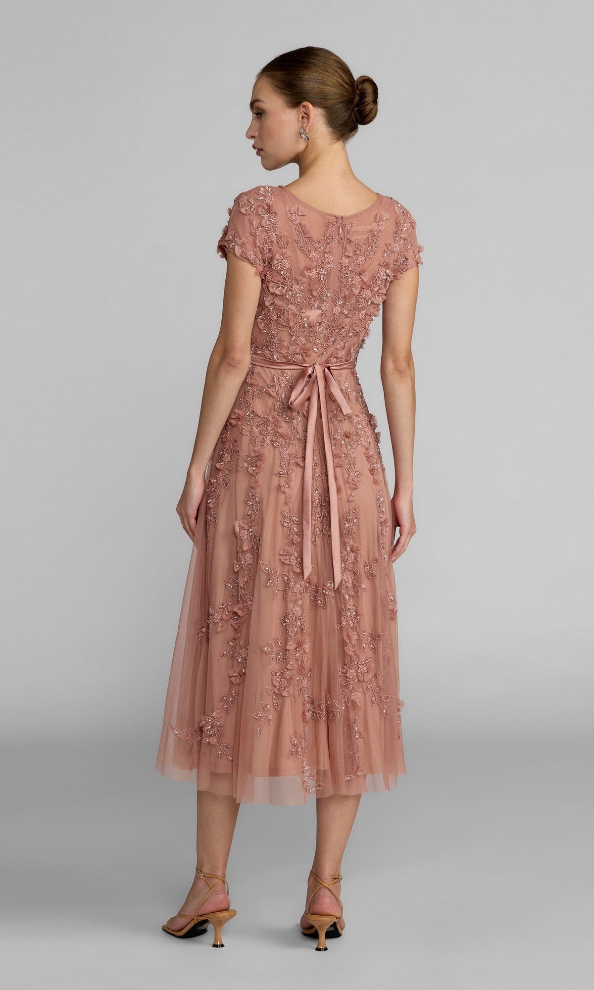 A woman faces away, wearing the Mac Duggal 94339 midi wedding guest dress in dusty pink with floral embroidery, ribbon waist tie, cap sleeves, and heeled sandals—ideal for a wedding reception.